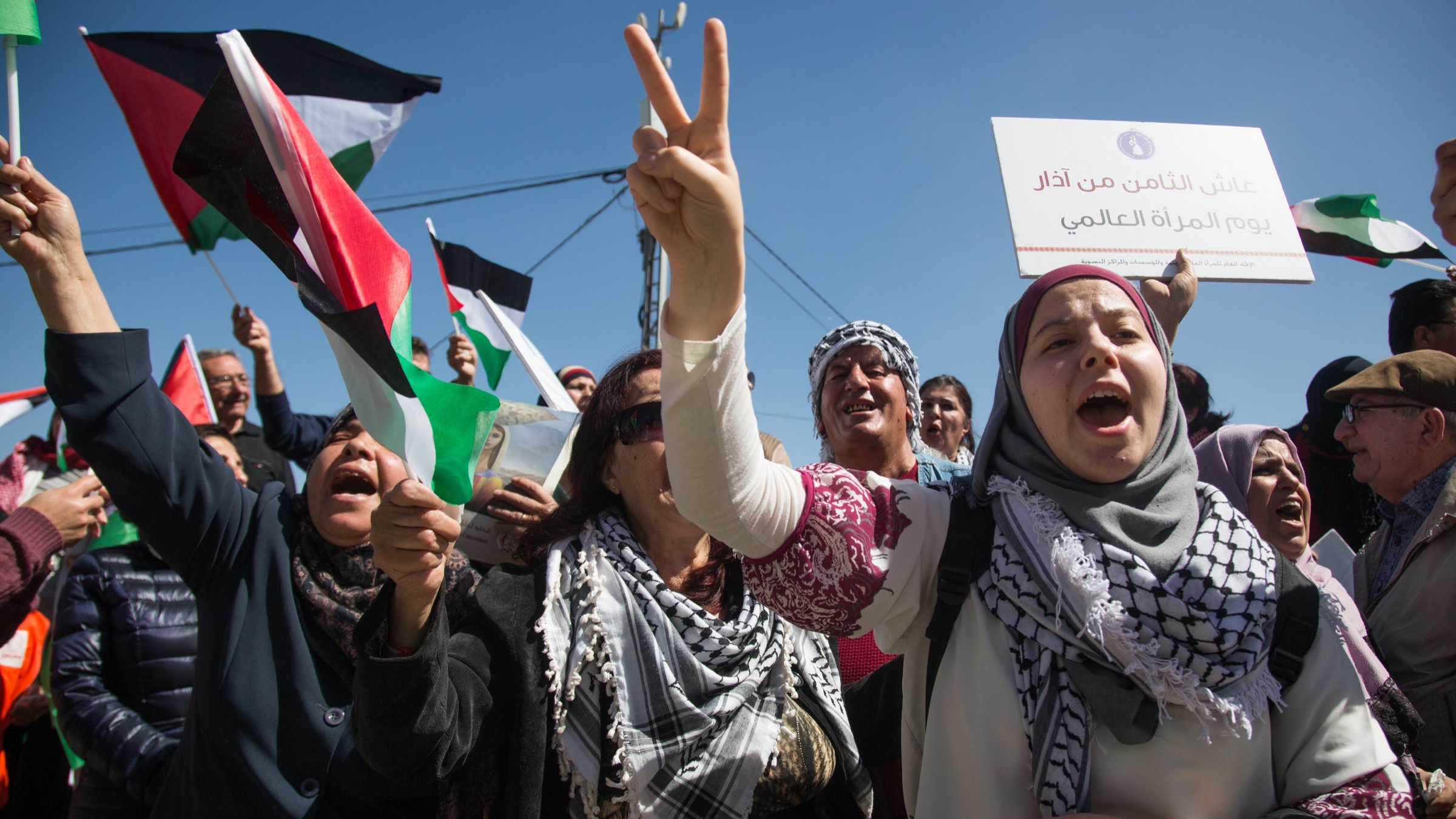 L'image montre une manifestation expressive, avec des personnes brandissant des drapeaux palestiniens et scandant des slogans. Les manifestants semblent passionnés et engagés, certains levant les bras en signe de victoire. Au premier plan, une femme en hijab tient un panneau et exprime son enthousiasme. L'arrière-plan est rempli de supporters, créant une atmosphère de solidarité et de détermination. La scène se déroule sous un ciel bleu, renforçant le sentiment d'énergie et de mobilisation.