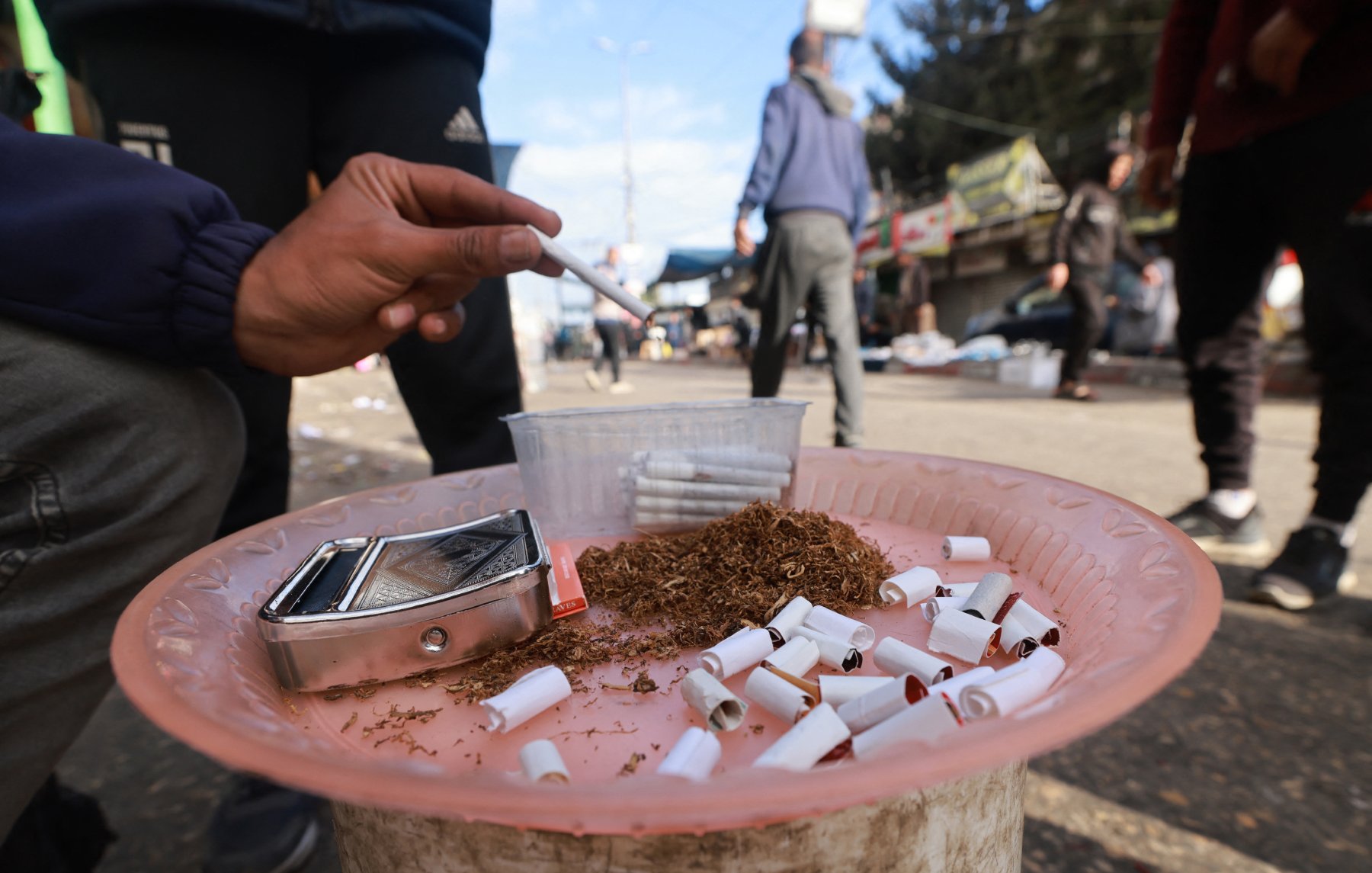 L'image montre une scène de rue avec une personne qui tient une cigarette à la main. Sur un plateau, on peut voir du tabac en vrac, des cigarettes déjà consommées et un petit récipient. En arrière-plan, on aperçoit des passants et des affiches sur des murs. L'ambiance semble animée et urbaine.