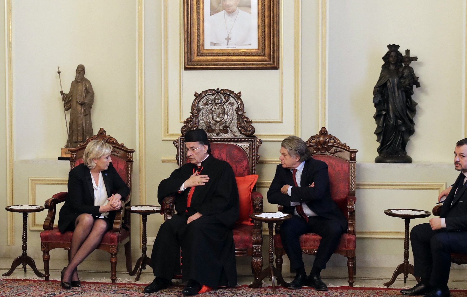 L'image montre une scène d'une réunion ou d'un entretien formel. Trois personnes sont assises dans des fauteuils ornés, au milieu d'une pièce décorée de manière élégante. L'une des personnes, portant une tenue ecclésiastique, semble être en train de parler, tandis que les autres l'écoutent attentivement. Les murs sont ornés de statues et d'œuvres d'art, ce qui suggère un environnement culturel et riche en histoire.