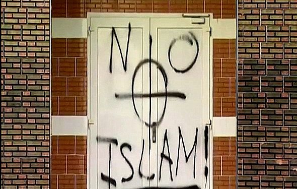 در این تصویر، درب یک ساختمان با رنگ سیاه نوشته شده است. در وسط درب علامت دایره‌ای با خطی عمودی و افقی وجود دارد که به همراه نوشته "NO ISLAM!" که به معنای "اسلام نه!" است، روی درب دیده می‌شود. این نوشته به روشنی یک پیام اعتراضی یا نفرت‌آمیز را منتقل می‌کند. زمینه دیوار نیز آجری و با رنگ‌های متفاوت است.