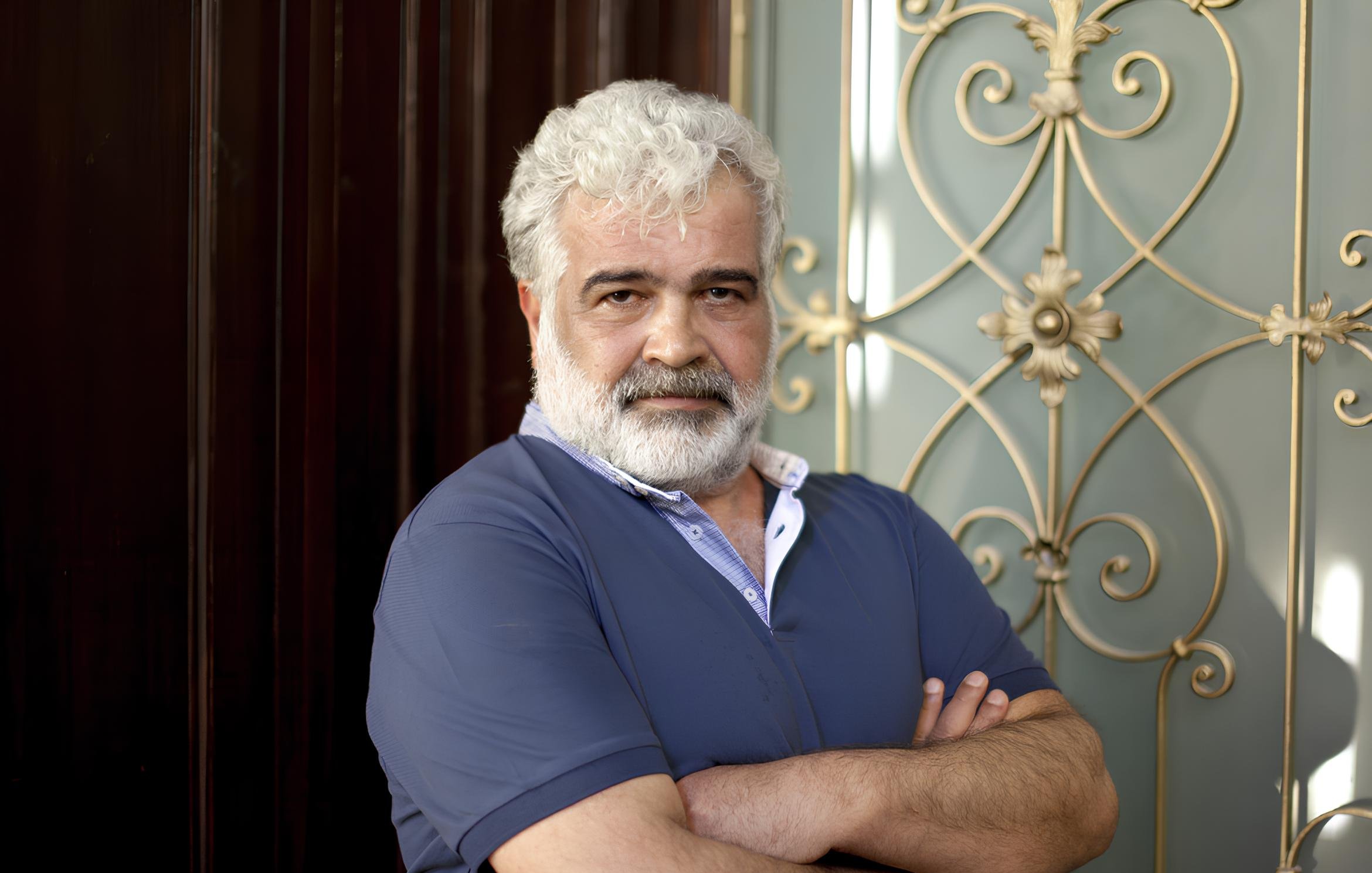 In questa immagine, si vede un uomo maturo con capelli bianchi e una barba folta. Indossa una maglietta blu e ha un'espressione seria, con le braccia incrociate. Sullo sfondo, ci sono elementi decorativi di metallo che aggiungono un tocco elegante all'ambiente. La luce mette in risalto i dettagli del suo viso e dell'abbigliamento.