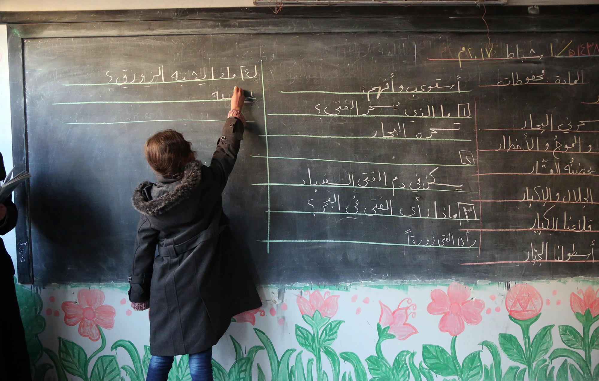 The image depicts a classroom scene where a student is writing on a blackboard. The blackboard has lines of text in Arabic, while the bottom of the board is decorated with colorful flower drawings. The student, wearing a coat, is focused on writing, and there are other individuals in the background, potentially teachers or classmates. The setting appears to be an educational environment.