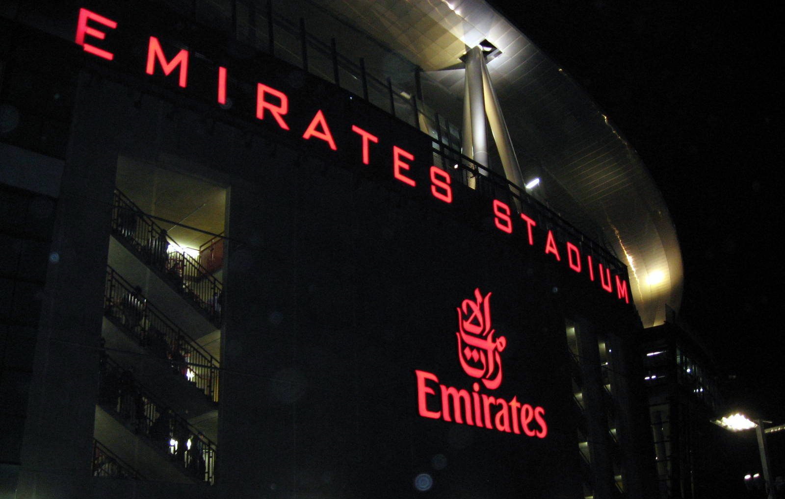 L'image montre l'extérieur de l'Emirates Stadium, qui est la maison du club de football Arsenal, à Londres. Le bâtiment est illuminé, avec le nom "EMIRATES STADIUM" en lettres rouges lumineuses bien visibles. On peut également voir le logo d'Emirates, une compagnie aérienne, mis en évidence. L'ambiance semble animée, suggérant qu'il y a probablement un événement ou un match en cours, avec des personnes rassemblées autour de la façade du stade.