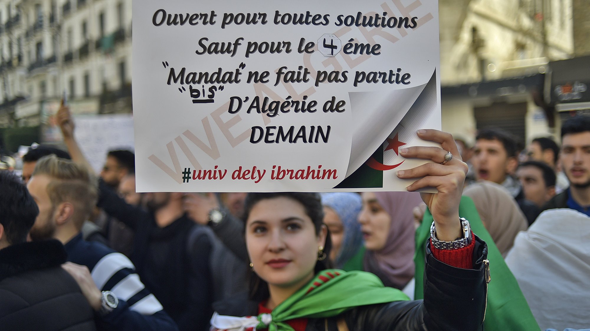 L'image montre une manifestation, où une jeune femme tient une pancarte. La pancarte indique qu'elle est ouverte à toutes solutions, sauf pour le 4ème mandat, affirmant que le mandat ne fait pas partie de l'Algérie de demain. La femme est entourée d'autres manifestants et porte un drapeau. L'atmosphère semble engagée et déterminée, reflétant des revendications politiques.