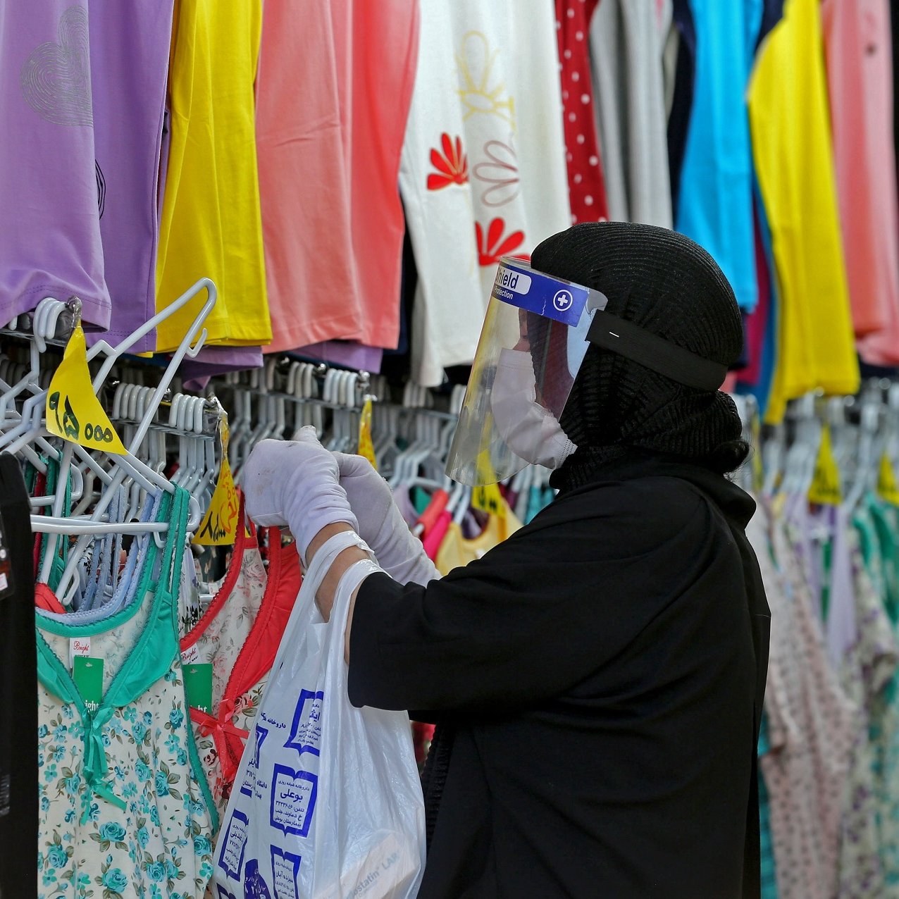 La imagen muestra a una persona en una tienda de ropa. Está usando una mascarilla y un protector facial. La persona está revisando prendas de vestir colgadas en perchas, que son de diversos colores y estilos. Se pueden ver camisetas y pantalones en el fondo, mientras que tiene una bolsa en la mano. La escena refleja un ambiente de compra de ropa, probablemente en un contexto donde se aplican medidas de seguridad relacionadas con la salud.