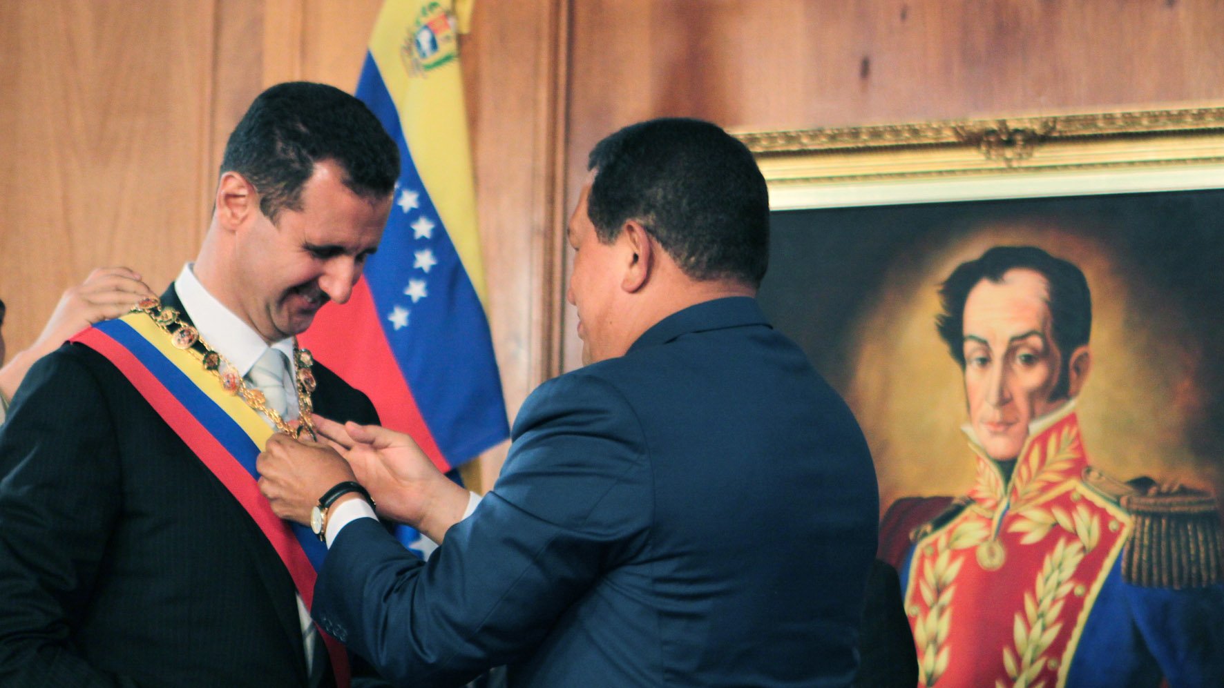 L'image montre une cérémonie officielle où un homme, probablement un leader politique, reçoit un insigne ou un collier de la part d'un autre homme. Le premier homme est vêtu d'un costume noir et porte une écharpe, tandis que le second, habillé en bleu, est en train de lui remettre l'insigne autour du cou. En arrière-plan, on aperçoit un portrait d'une figure historique, ainsi que des drapeaux, ce qui suggère un contexte politique et national.