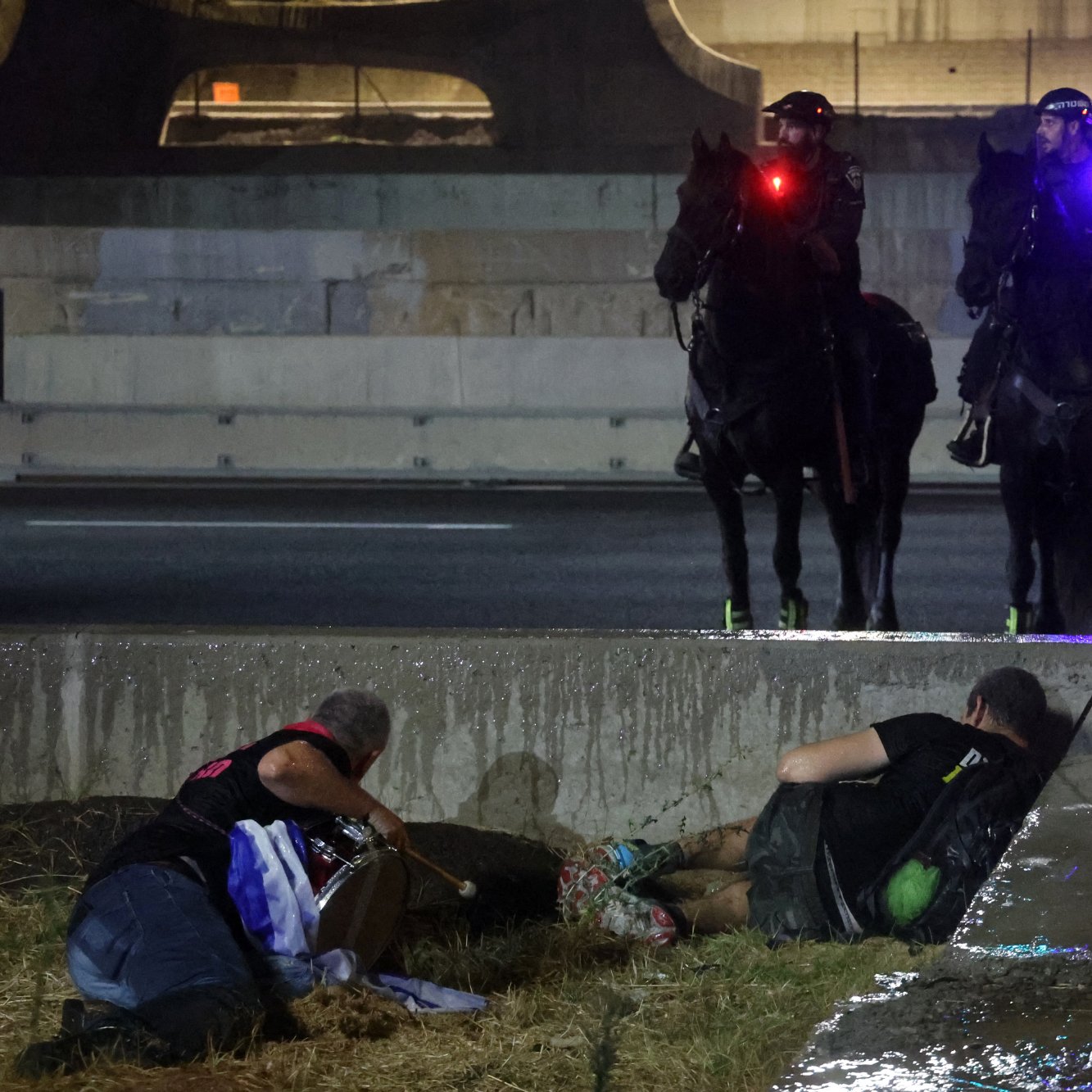 تصویر نشان‌دهنده‌ی دو نفر است که در حال استراحت یا پنهان شدن در کنار یک جاده هستند. در پس‌زمینه، دو پلیس با اسب‌های خود دیده می‌شوند که چراغ‌های قرمز و آبی بر روی سر اسب‌ها روشن است. همچنین، پرچم اسرائیل در نزدیکی آن‌ها قرار دارد و نشان‌دهنده‌ی یک وضعیت تنش‌آلود یا اعتراض است. زمین مرطوب به نظر می‌رسد و جو تصویر حس درگیری یا تنش را منتقل می‌کند.