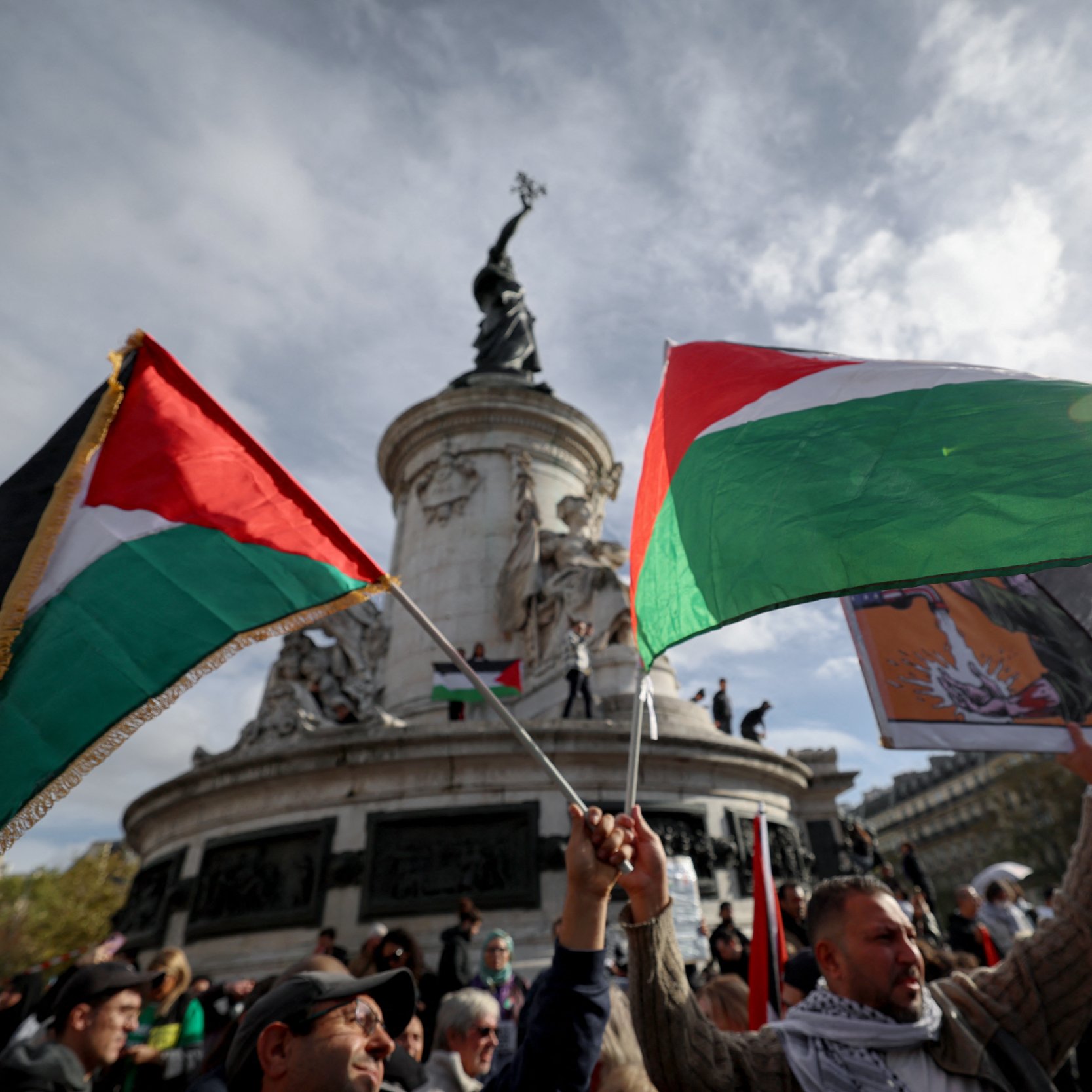 L'image montre une scène de manifestation avec des participants brandissant des drapeaux palestiniens. En arrière-plan, on peut voir un monument, probablement une statue, qui est entouré d'un ciel nuageux. Les personnes présentes semblent exprimant des sentiments forts, et l'atmosphère générale est celle d'un rassemblement public engagé et solennel.