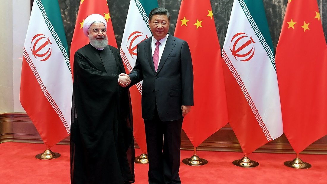 The image depicts two men shaking hands, standing in front of flags of Iran and China. The man on the left is wearing a black robe and white turban, representing Iran, while the man on the right is dressed in a suit. They appear to be in a formal setting, likely symbolizing a diplomatic meeting between the two countries. The background features a red carpet and prominently displayed national flags.