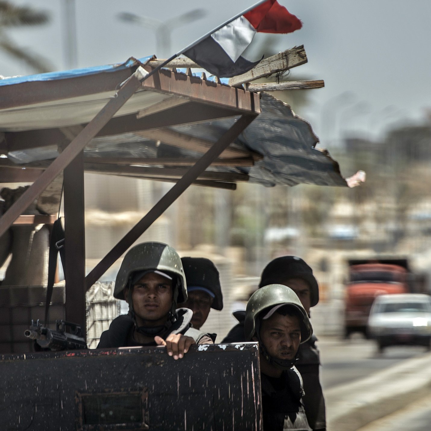الصورة تُظهر مجموعة من الجنود أو رجال الأمن، يرتدون بدلات عسكرية وخوذ، ويبدو أنهم على أهبة الاستعداد. يقفون خلف حاجز أو نقطة تفتيش، بينما يُمكن رؤية العلم المصري يُرفرف في الخلفية. المشهد يتضمن أيضًا بعض التفاصيل عن البيئة المحيطة، مثل الشوارع والمباني. الأجواء تبدو مشحونة، مما يوحي بوجود حالة تأهب أو تواجد أمني مكثف.