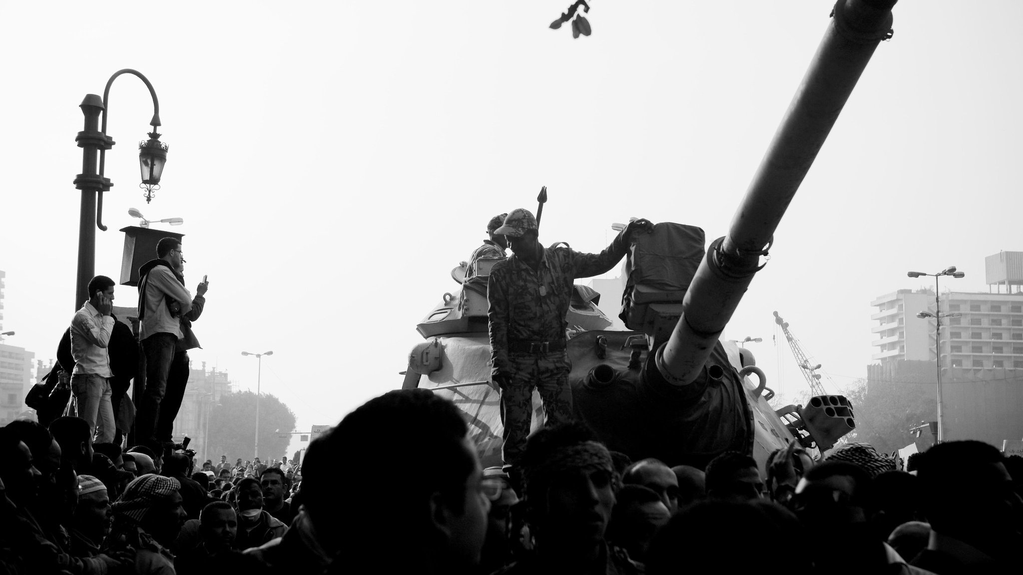 تظهر الصورة مشهداً من مظاهرة أو حدث احتجاجي، حيث يوجد عدد كبير من الناس يتجمعون حول دبابة. أحد الجنود يقف فوق الدبابة ويرفع يدَه كما لو كان يحمل شيئاً أو يجذب الانتباه. الأجواء تشير إلى توتر أو حالة من الحماس بين الحشود. الصورة تُظهر تبايناً كبيراً بين الأضواء والظلال، مما يضفي عليها طابعاً دراماتيكياً.