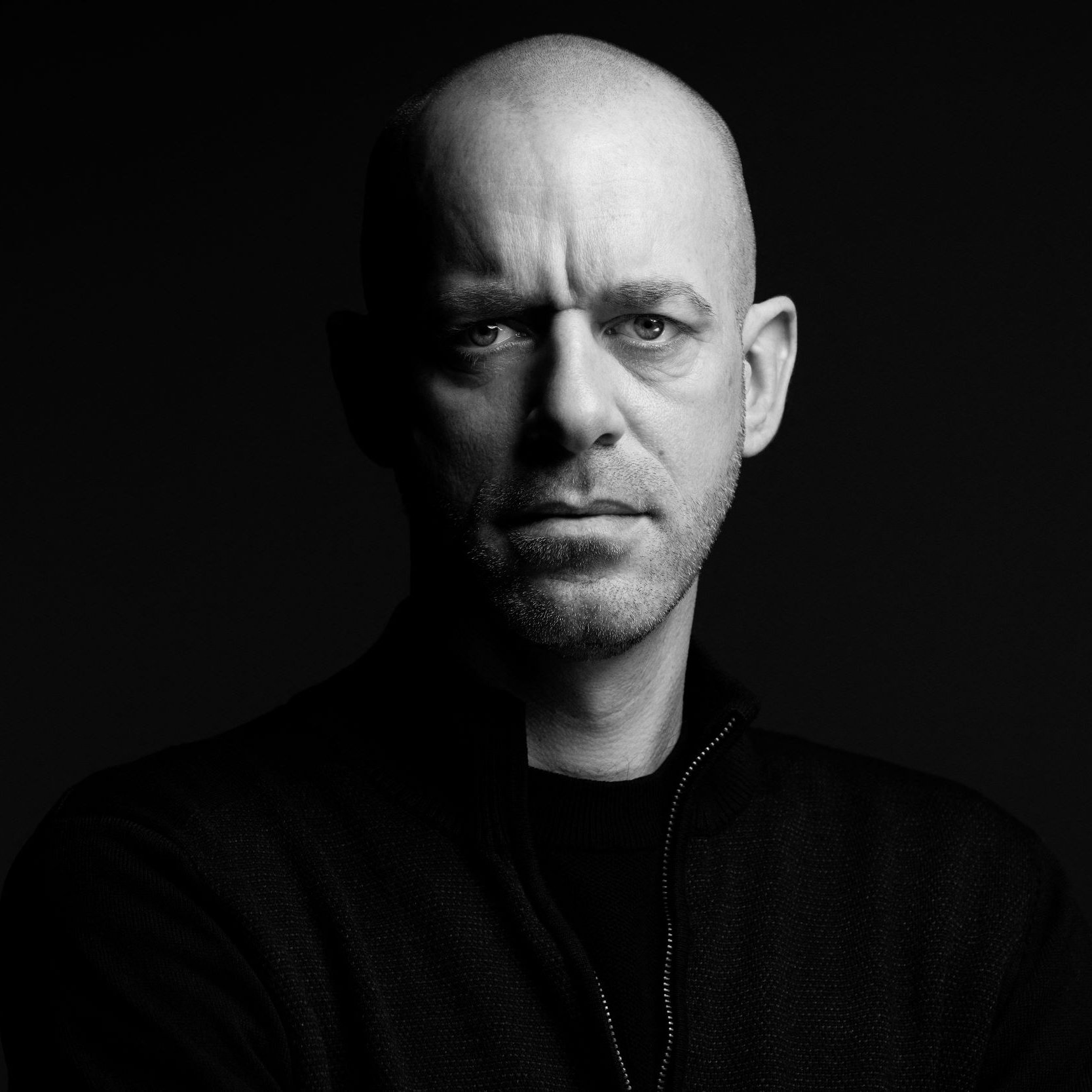 La imagen muestra a un hombre con cabeza rapada y una expresión seria. Está vestido con una prenda de color oscuro y parece estar mirando hacia la cámara. El fondo es negro, lo que resalta su figura y la iluminación hace que su rostro y gestos sean más definidos, creando un contraste dramático. La atmósfera de la imagen es intensa y poderosa.