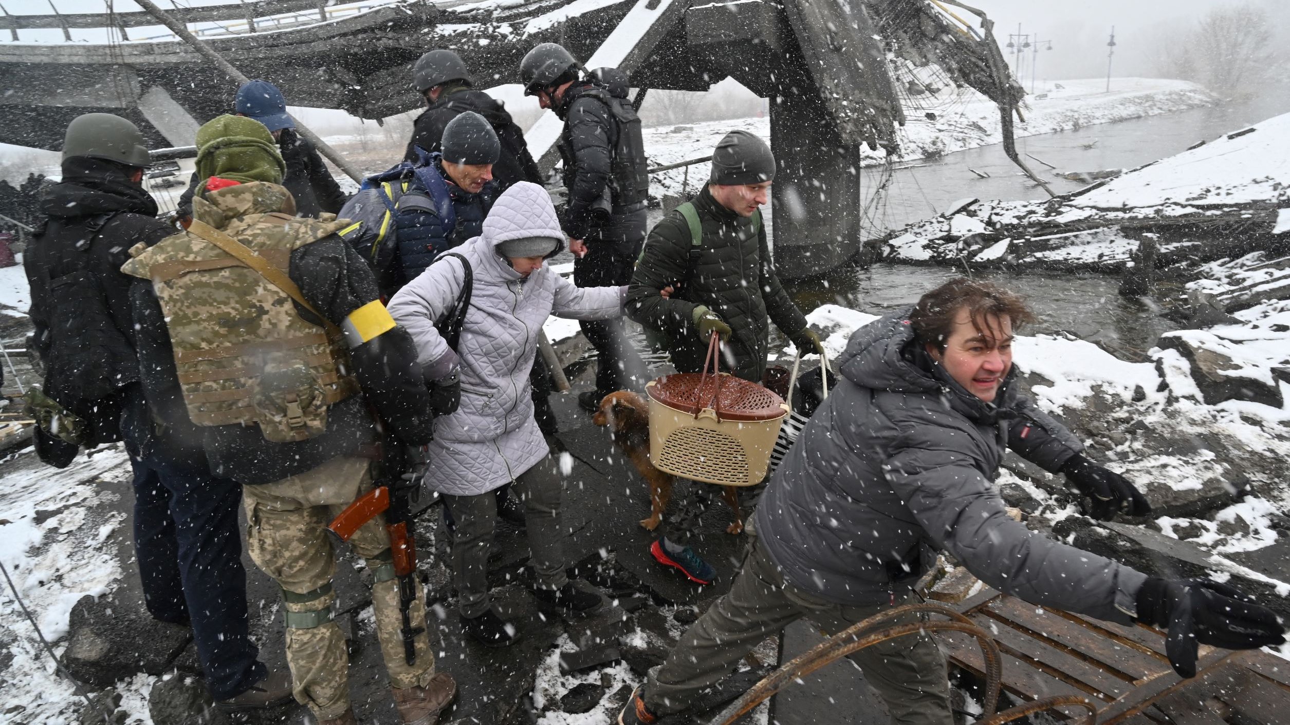 The image depicts a group of people navigating through a damaged area, likely following a disaster or conflict. They are surrounded by debris and snow, suggesting challenging weather conditions. Some individuals appear to be assisting others, while one person is pulling or reaching for something. The scene conveys a sense of urgency and struggle in a harsh environment, highlighting the effects of destruction and the resilience of those affected.
