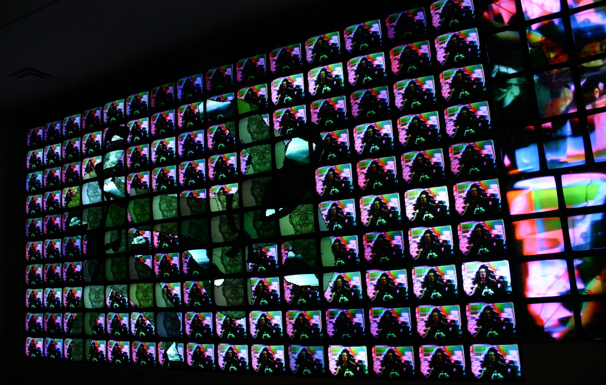 L'image présente une installation artistique composée de nombreux écrans alignés de manière à former un tableau visuel. Chaque écran affiche des images colorées, vibrantes et abstraites, créant un effet d'ensemble dynamique. Les couleurs se mélangent et se chevauchent, ajoutant une sensation de mouvement. La composition semble jouer avec la lumière et les contrastes, attirant l'œil vers des motifs diversifiés. L'ensemble évoque une immersion dans une expérience numérique moderne et artistique.