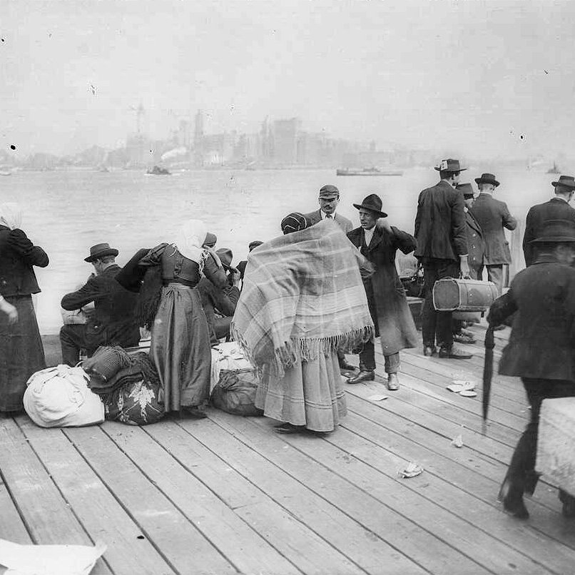 L'image montre un groupe de personnes rassemblées sur un quai, probablement à une époque antérieure. On peut voir des individus portant des habits typiques de l'époque, dont des chapeaux, et certains sont emmitouflés dans des couvertures. Ils semblent s'affairer autour de bagages et de marchandises, évoquant des thèmes de migration ou de transit. En arrière-plan, on aperçoit des bâtiments ainsi que des bateaux sur l'eau, suggérant un port animé. L'atmosphère semble à la fois chargée et historique.