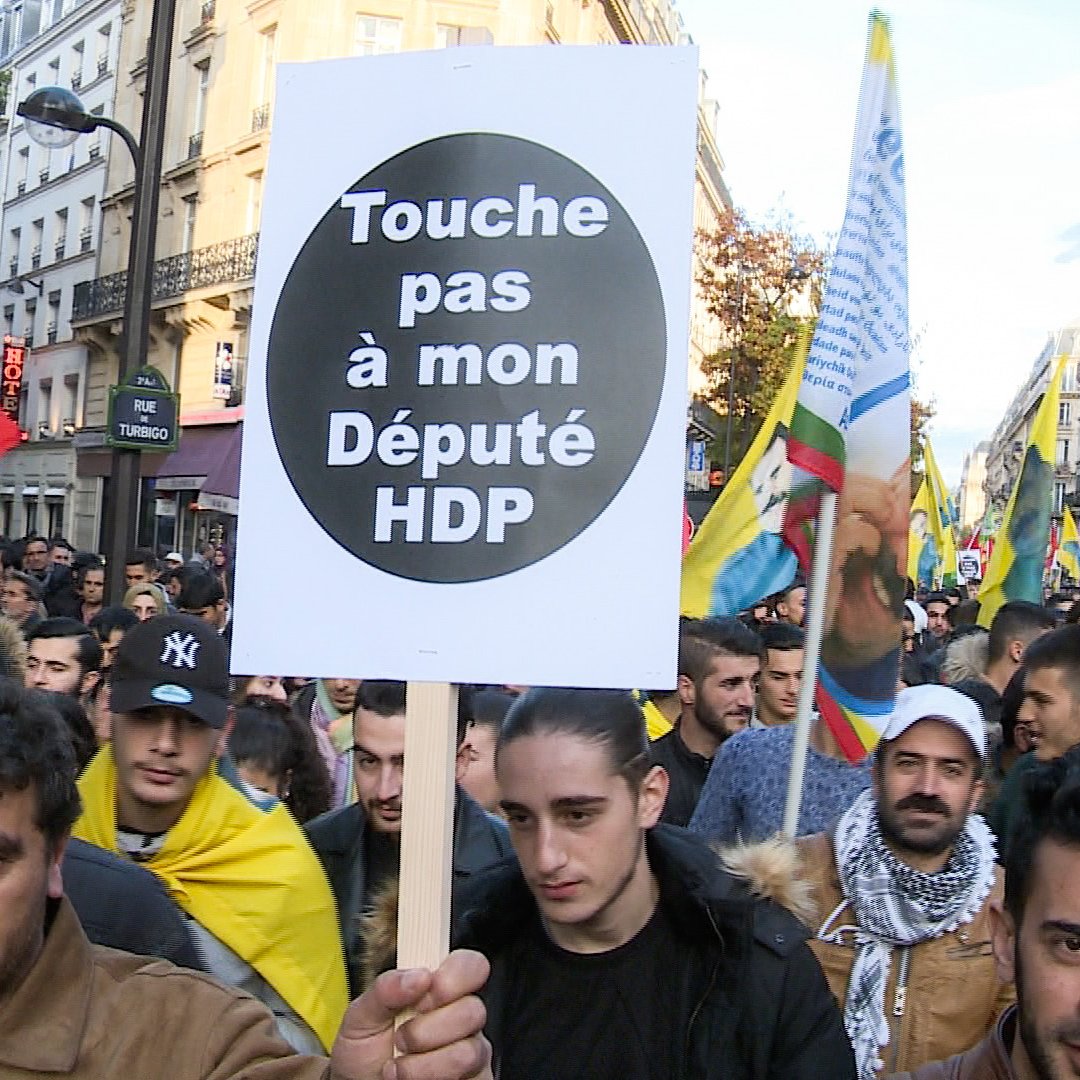 L'image montre une manifestation de foule dans une rue, où des personnes tiennent des pancartes et des drapeaux. Au premier plan, un manifestant porte une grande pancarte avec le message "Touche pas à mon Député HDP". L'ambiance semble être celle d'une protestation politique, impliquant probablement des questions liées au parti HDP (Parti démocratique des peuples) en Turquie. Les participants semblent engagés et déterminés.