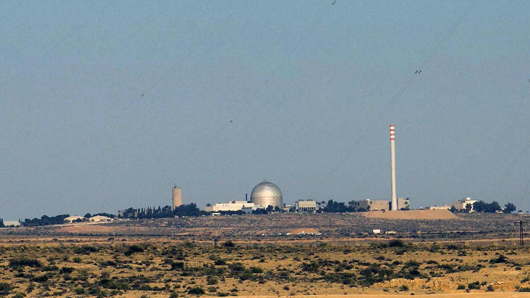 L'image montre un paysage désertique avec des bâtiments en arrière-plan. Au centre, on peut voir un dôme argenté, probablement un réacteur ou une installation scientifique. À côté, il y a une cheminée de grande taille et d'autres structures de différentes hauteurs, suggérant qu'il s'agit d'une installation industrielle ou scientifique. Le ciel est clair et il n'y a pas de nuages. Le terrain autour est aride, avec peu de végétation.