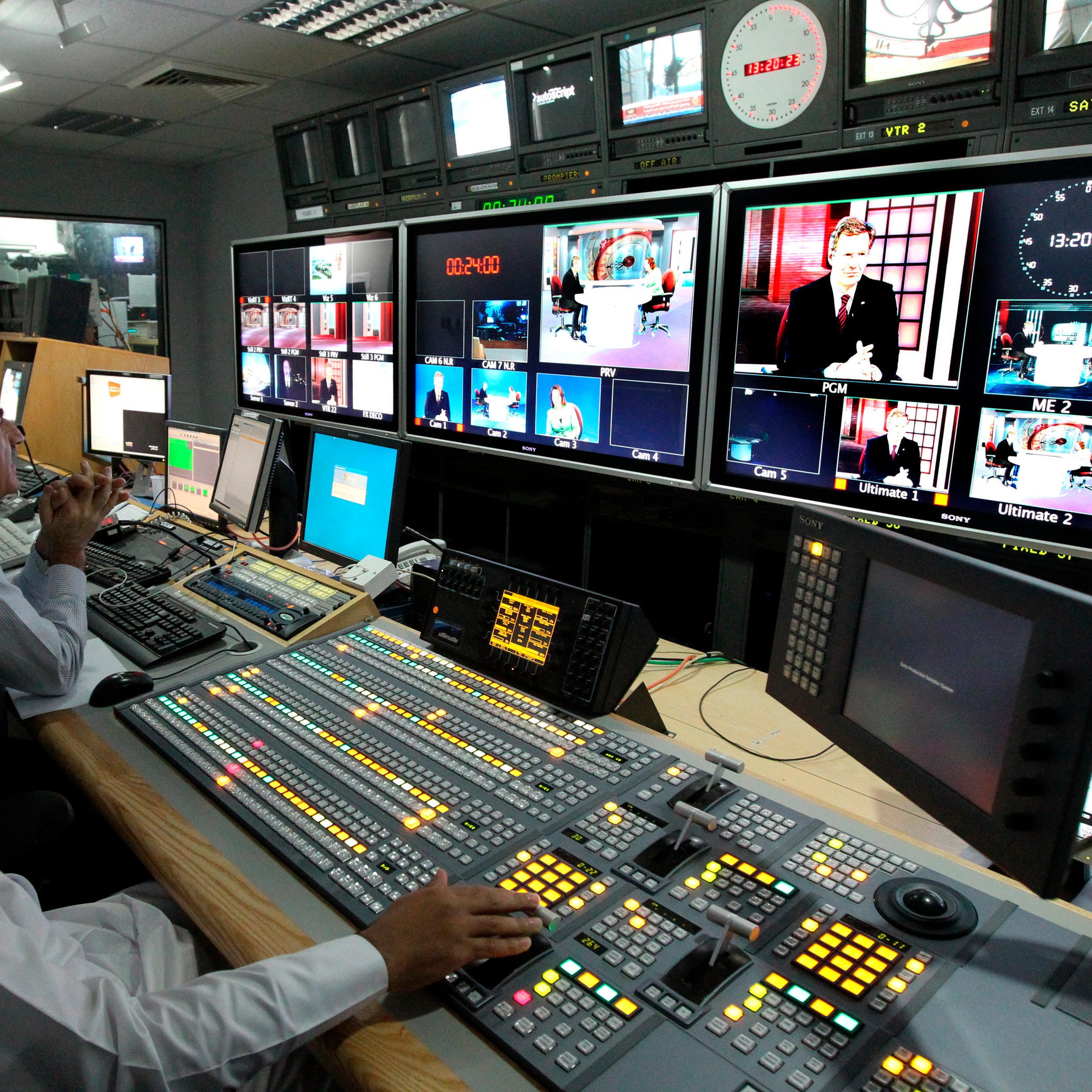 تظهر الصورة غرفة تحكم تلفزيونية. في المقدمة، يوجد شخصان يجلسان أمام معدات التحكم، مع وجود مجموعة من الأزرار والشاشات! أحدهم يبدو مركزًا على العمل، بينما الآخر يحمل هاتفًا ويبدو مشغولًا. في الخلف، يقف شخص ثالث يتابع الشاشات. تبرز من خلفهم مجموعة من الشاشات الكبيرة تعرض برامج وأخبار مختلفة، مع وجود ساعة تُظهر الوقت. الإضاءة في المكان طبيعية وتضفي أجواء احترافية.