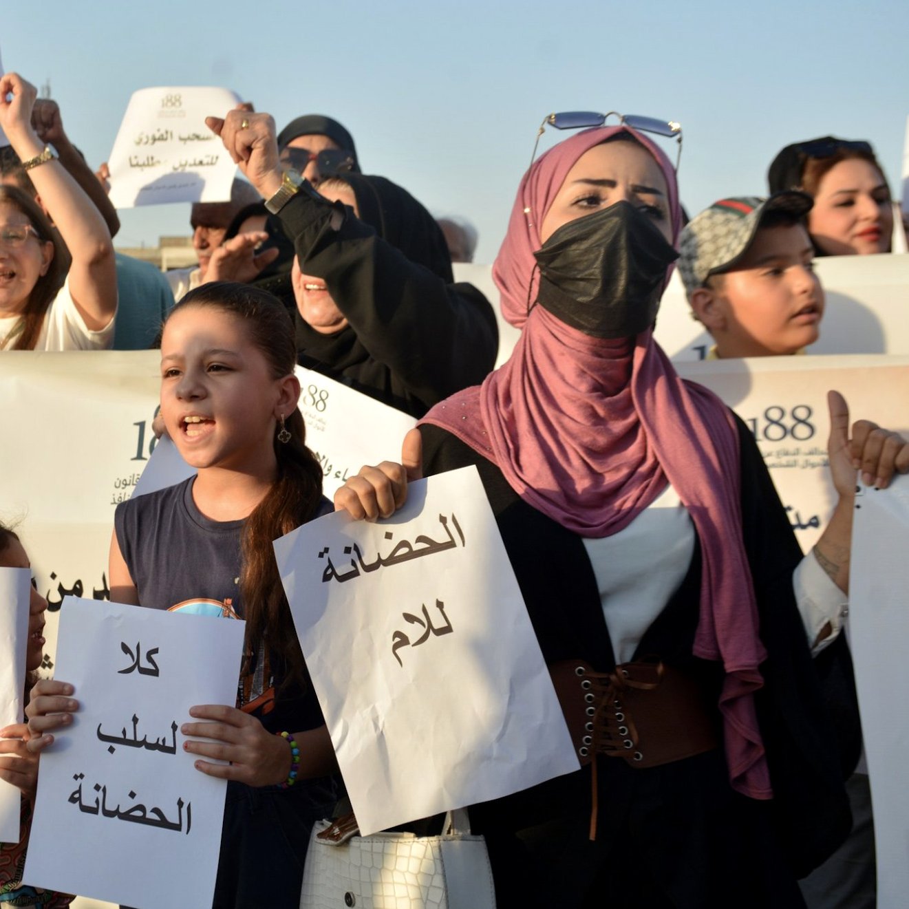 در این تصویر، گروهی از افراد در حال برگزاری یک تجمع اعتراضی هستند. زنان و مردان در این جمع حضور دارند و برخی از آن‌ها پلاکاردهایی با نوشته‌هایی در دست دارند. یک دختر بچه در جلوتر ایستاده و به وضوح در حال سخنرانی است. بیشتر افراد از لباس‌های متنوعی استفاده کرده‌اند و به نظر می‌رسد که همه آنها به یک مسئله اجتماعی یا سیاسی اهمیت زیادی می‌دهند. جو عمومی تصویر نشان‌دهنده شور و هیجان در این تجمع است.