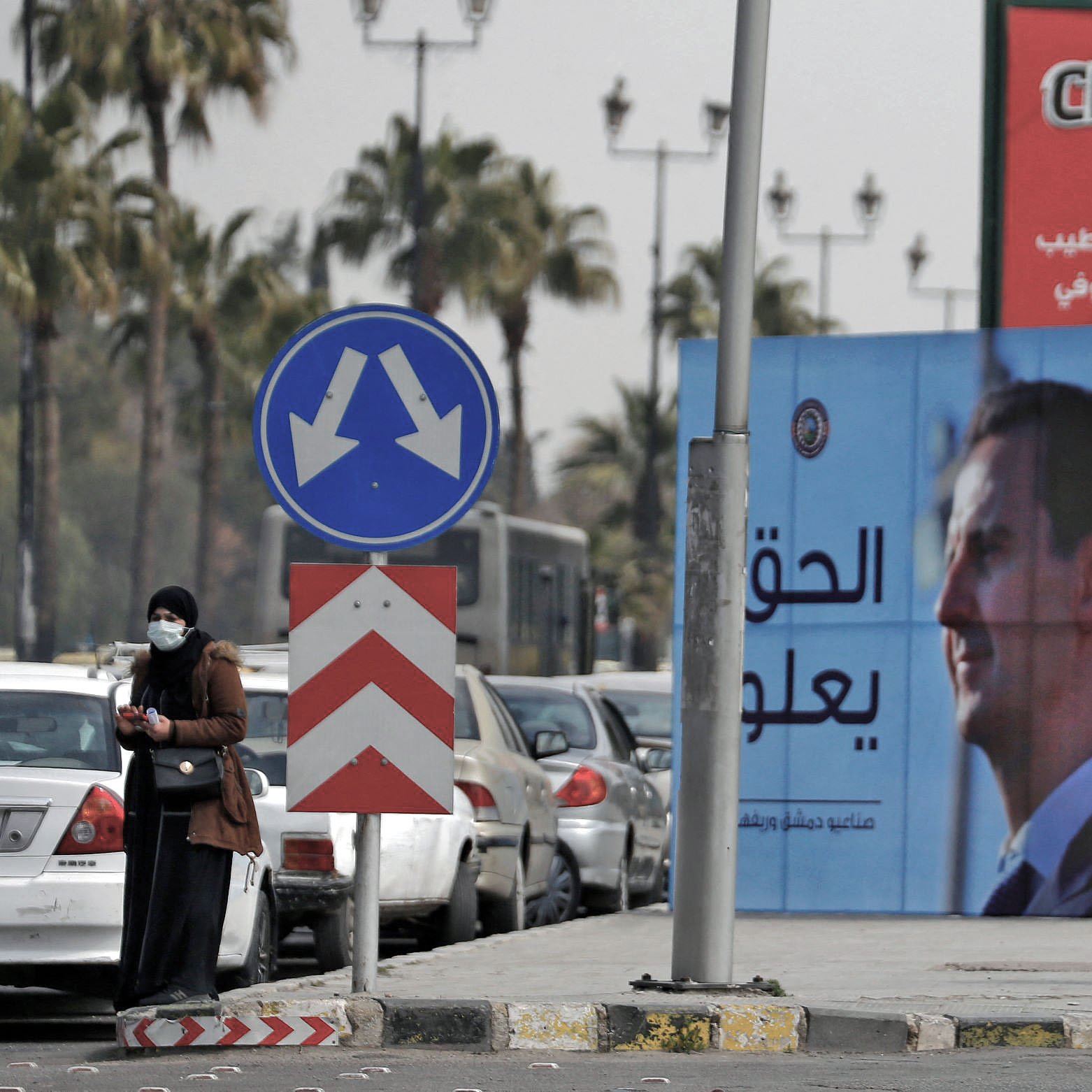 در این تصویر، خیابانی مطرح شده و در آن دو نفر در کنار پیاده‌رو ایستاده‌اند. در پس‌زمینه، چندین خودرو پارک شده و همچنین یک بیلبورد بزرگ با تصاویر و متنی به زبان عربی دیده می‌شود. این بیلبورد به احتمال زیاد به مقامات و موضوعات سیاسی اشاره دارد. تبلیغاتی برای یک برند معروف تنقلات نیز در بالای تصویر دیده می‌شود. در مجموع، تصویر حال و هوای شهری را به نمایش می‌گذارد.