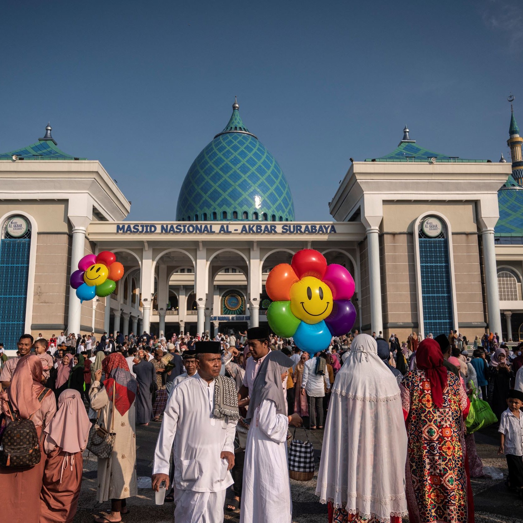 The image depicts a large crowd gathered outside the National Mosque of Al-Akbar in Surabaya, Indonesia. The mosque features a prominent architectural style with a blue and green patterned dome. In the foreground, people of various ages and attire are seen, many wearing traditional Islamic clothing. Some individuals are holding colorful balloons with smiling faces, adding a festive atmosphere to the scene. The sky is clear and blue, suggesting a bright and sunny day.