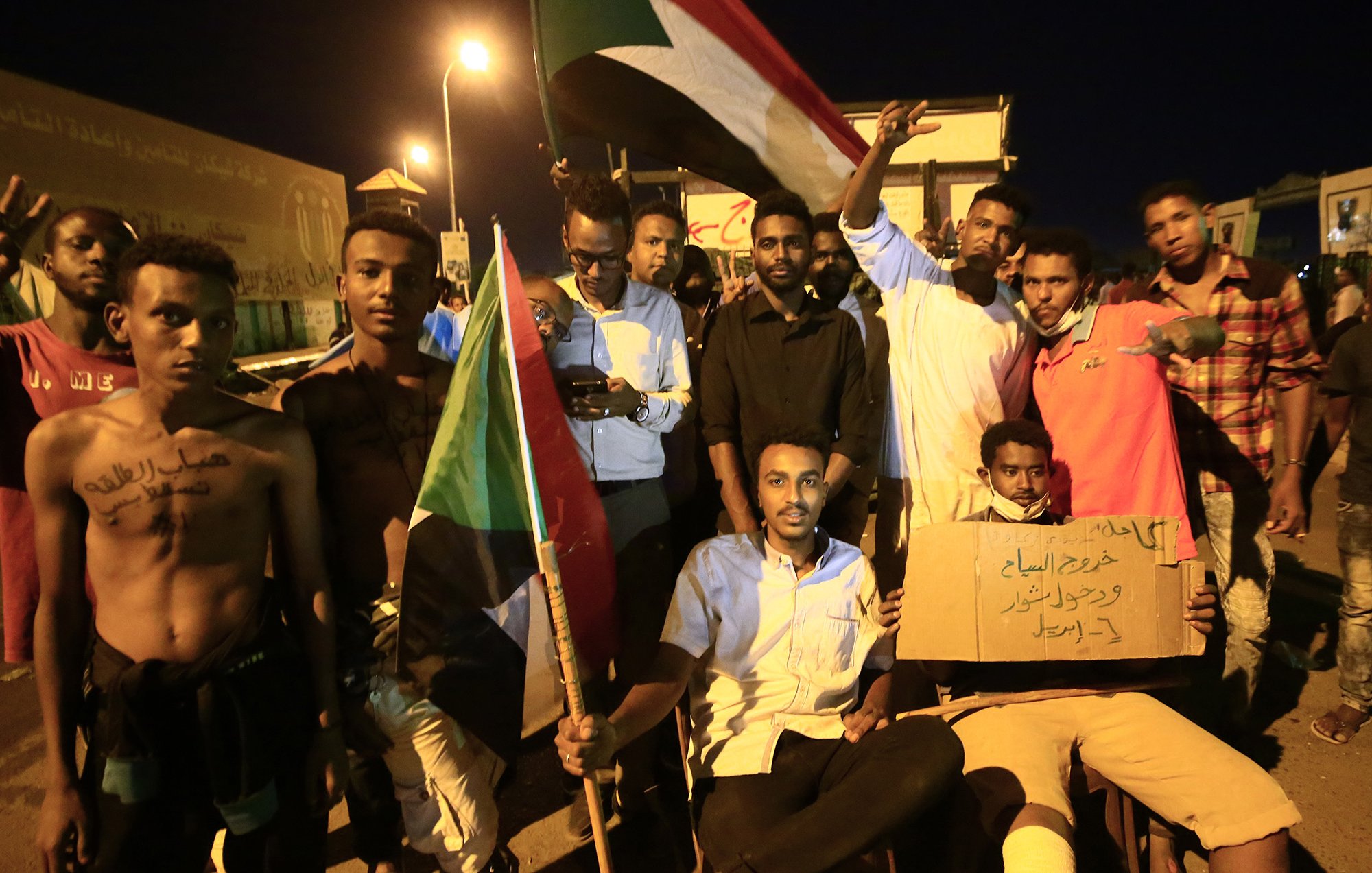 L'image montre un groupe de manifestants soudanais rassemblés lors d'une nuit, tenant des drapeaux et des pancartes. Certains d'entre eux sont debout, affichant des signes de solidarité avec les bras levés, tandis que d'autres sont assis, tenant des panneaux de revendication. L'atmosphère semble énergique et déterminée, incarnant un esprit de protestation et de demande de changement. Les visages des participants expriment diverses émotions, allant de la détermination à l'espoir.