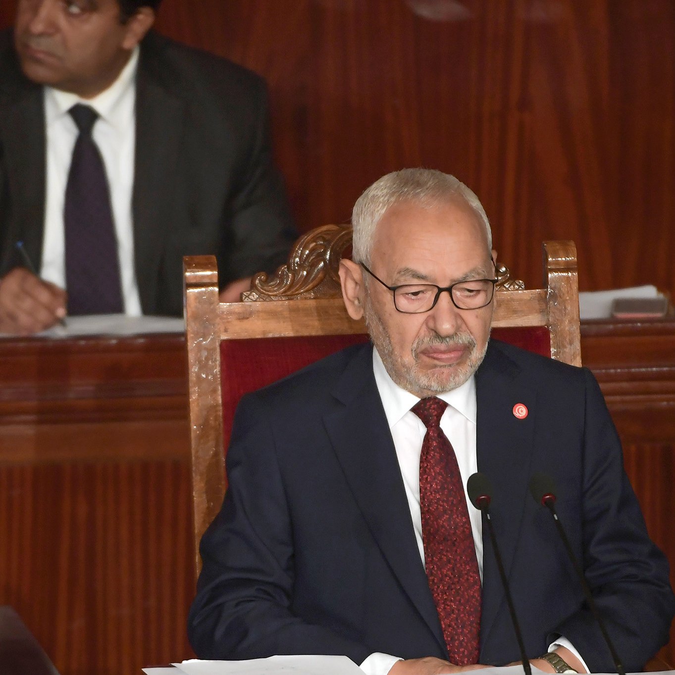 The image shows a man sitting at a podium in a formal setting, likely a legislative or parliamentary assembly. He is wearing a dark suit and glasses, and appears to be speaking or presenting. In the background, another man is seated and observing, dressed in a suit and tie. The environment has a wooden backdrop that contributes to a serious and official atmosphere.