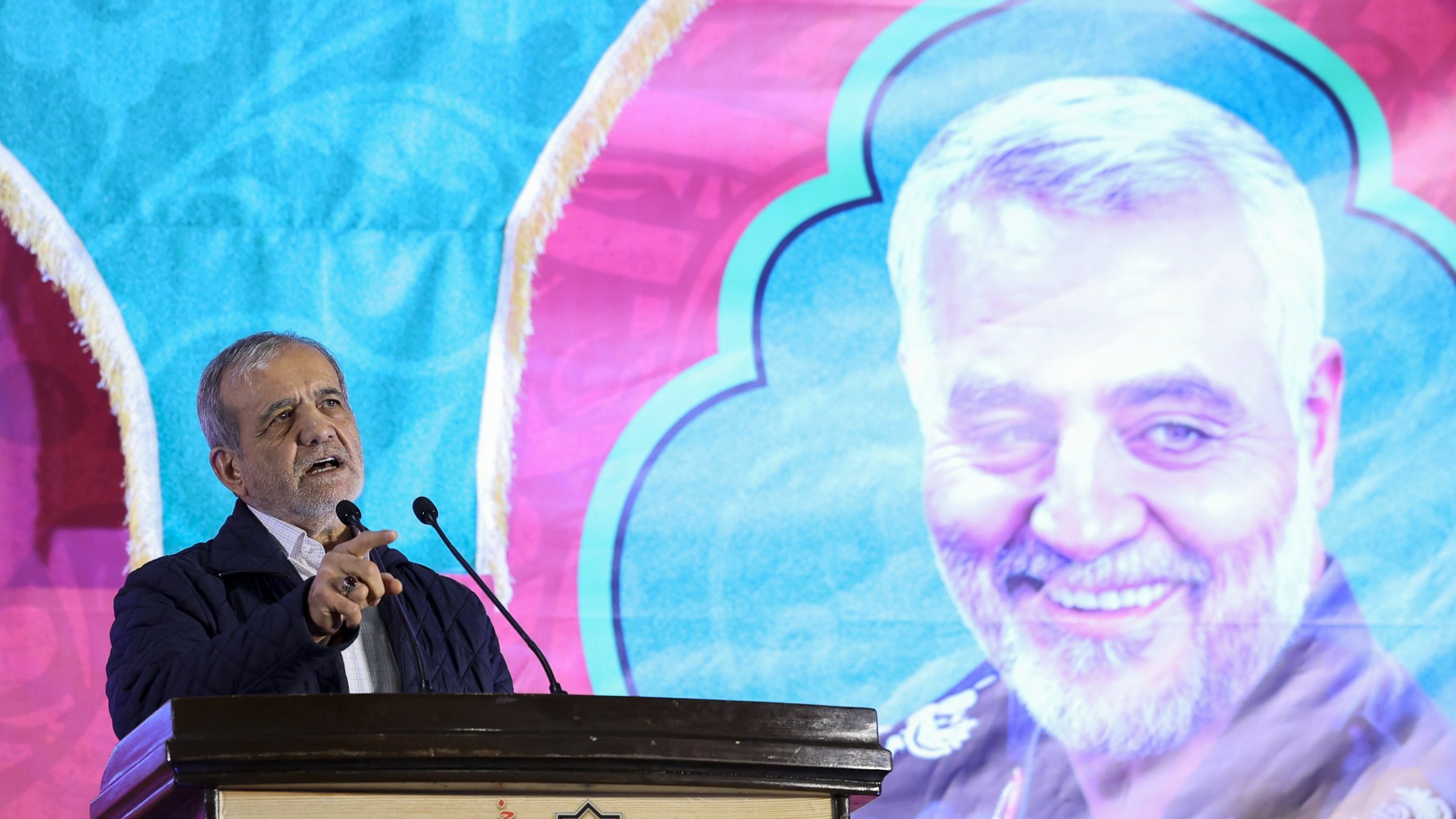 The image features a man speaking at a podium, with a colorful backdrop that includes a large portrait of another individual smiling. The background has a decorative design, predominantly featuring blue and pink colors. The speaker appears animated and engaged, and the setting suggests it may be a formal event or speech, possibly commemorating or honoring the person depicted in the portrait.