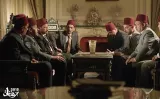 L'image montre un groupe d'hommes assis dans une pièce élégamment meublée. Ils portent des fez rouges, ce qui indique un contexte culturel spécifique. Les hommes semblent engagés dans une discussion sérieuse, et la disposition de leurs corps suggère un débat ou une réunion importante. L'ambiance de la pièce est raffinée, avec des éléments de décor qui renforcent un cadre formel. Leurs expressions faciales indiquent un mélange de concentration et de tension.