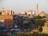 L'image montre une vue d'une ville avec un mélange d'architecture moderne et traditionnelle. On peut voir plusieurs bâtiments de différentes hauteurs, avec des toits en tuiles et en béton. À l'arrière-plan, il y a un minaret et un grand dôme, ce qui suggère la présence d'une mosquée. Le ciel est clair, indiquant probablement une matinée ou un après-midi ensoleillé. De plus, plusieurs antennes parabolique sont visibles sur les toits, ce qui reflète une urbanisation moderne.