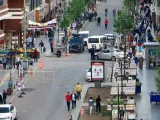 L'image montre une rue animée, avec de nombreuses personnes marchant sur le trottoir et dans la rue. On voit des véhicules stationnés et d'autres en circulation. La scène est bordée de magasins et de kiosques, créant une atmosphère vivante. Des arbres et des décorations peuvent également être visibles, ajoutant une touche de verdure et de couleur. Au loin, on aperçoit des gens engagés dans diverses activités, ce qui contribue à l'animation de l'environnement urbain.