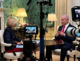 Deux personnes en interview, un homme en costume et une femme, devant un décor élégant.