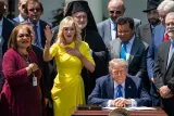 Un homme assis, signe un document, entouré de personnes en arrière-plan, une femme en jaune.