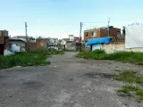 L'image montre un terrain vague ou une rue désaffectée, entourée de bâtiments en briques. Il y a une certaine végétation, avec de l'herbe qui pousse sur le sol poussiéreux. En arrière-plan, on aperçoit des structures en béton et des toits, certains recouverts de bâches bleues ou de toiles. Le ciel est clair, et l'ensemble de la scène dégage une impression de désolation et d'abandon.