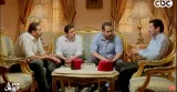 L'image montre quatre hommes assis autour d'une table dans un cadre intérieur décoré avec des rideaux dorés. Ils portent des chemises et semblent avoir une discussion animée. Deux des hommes portent un fez rouge sur la table. Le mobilier est élégant, avec un canapé de style classique et une table en verre au centre. L'ambiance semble conviviale et décontractée.