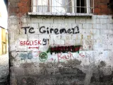 L'image montre un mur en briques avec des taches d'humidité et des signes d'usure. Sur le mur, on peut voir des graffitis, dont des mots et des symboles écrits en noir, rouge et vert. Certaines inscriptions semblent être des messages ou des tags, et le mur présente une texture irrégulière, typique des bâtiments anciens ou abandonnés. Le sol est constitué de pavés, indiquant un environnement urbain.