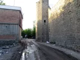 L'image montre une rue étroite et poussiéreuse, bordée de murs en pierre. À droite, on peut voir un ancien mur en pierre, probablement une partie d'une ancienne fortification. À gauche, il y a un bâtiment en briques avec une façade moins bien entretenue. La route est légèrement humide et semble avoir des traces de passage avec quelques déchets visibles sur le sol. En arrière-plan, on aperçoit des arbres, indiquant une certaine verdure dans les environs. L'atmosphère est calme et un peu délaissée.