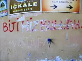 L'image montre un mur sur lequel sont peints des graffitis. L'inscription principale, en lettres rouges, dit "BIJÎ HIZBULLAH". Il y a également d'autres inscriptions, probablement de différentes couleurs, qui semblent moins lisibles. Le mur est entouré de panneaux publicitaires en turc pour des cafés et des petits-déjeuners. L'ensemble crée une ambiance urbaine et témoigne de l'usage de l'espace public pour exprimer des messages.