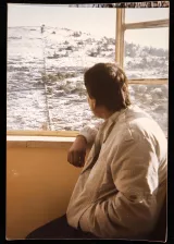 L'immagine mostra un uomo seduto accanto a una finestra, mentre guarda verso un paesaggio innevato. Indossa una giacca chiara e ha i capelli scuri. Fuori dalla finestra si possono vedere colline coperte di neve, suggerendo un'atmosfera tranquilla e invernale. La luce sembra calda, creando un contrasto con il freddo esterno. La scena trasmette un senso di riflessione e contemplazione.