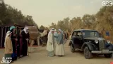 L'image semble représenter une scène se déroulant dans un environnement désertique ou villageois. On peut y voir plusieurs personnages vêtus de vêtements traditionnels, discutant près d'une voiture ancienne. À proximité, un chameau est visible, stable, et des tentes typiques de la région sont installées en arrière-plan. L'ambiance évoque un mélange de traditions anciennes et d'éléments modernes, comme la voiture. Les personnages semblent engagés dans une interaction, suggérant un moment de rencontre ou de négociation.