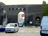 L'image montre un ancien portail en pierre, peut-être une porte d'une ville ou d'une fortification. Au premier plan, il y a des véhicules stationnés sur une surface pavée. Des câbles électriques sont visibles au-dessus, et on aperçoit des bâtiments modernes en arrière-plan. Un panneau rouge avec un symbole identifiable est accroché sur le portail. Des éléments de nature, comme des arbres, ajoutent une touche de verdure à l'ensemble.