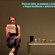 Una donna legge appunti davanti a un tavolo con piante e oggetti, su uno sfondo neutro.