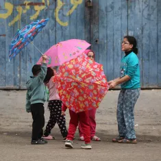 L'immagine mostra un gruppo di bambini che giocano insieme in strada, mentre tengono in mano degli ombrelli colorati. I bambini indossano abiti semplici e variopinti, e sembrano divertirsi sotto un muro blu con scritte. La scena trasmette gioia e convivialità.