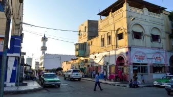 L'image montre une scène de rue animée dans une ville. On peut voir des bâtiments aux façades jaunes et blanches, typiques de l'architecture locale. À gauche, il y a des boutiques et un restaurant avec une enseigne bien visible. Sur la droite, un homme traverse la rue, entouré de véhicules stationnés et circulant. En arrière-plan, un minaret se dresse, indiquant la présence d'une mosquée. L'atmosphère est vivante, avec des passants et des couleurs vives qui caractérisent cet environnement urbain.