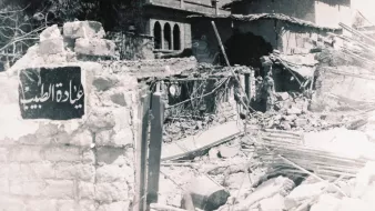 L'image montre une scène de destruction, avec des bâtiments en ruines. On peut apercevoir un mur effondré, laissant entrevoir des débris et des structures endommagées. À côté des débris, il y a une plaque signalétique en arabe qui semble indiquer un lieu ou un bâtiment. L'atmosphère générale de la photo suggère un environnement dévasté, probablement à la suite d'un conflit ou d'un désastre.