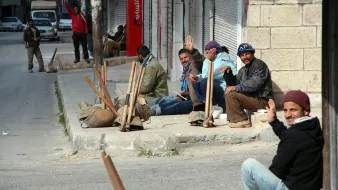 The image depicts a group of men sitting on a sidewalk in an urban setting. They appear to be relaxed, some waving at the camera, and are dressed in casual clothing. The street is relatively quiet, with a few vehicles in the background. There are also some construction tools and materials scattered around, suggesting they may be waiting for work or gathering. The environment looks somewhat desolate, with shuttered storefronts and a hint of a dusty road.