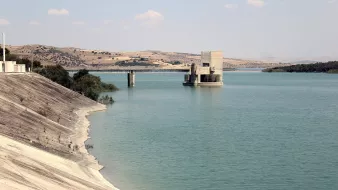 L'image montre un paysage calme avec un grand plan d'eau, probablement un réservoir ou un lac. Sur la gauche, la rive est recouverte d'une surface lisse et légèrement inclinée, tandis qu'à droite, on peut voir une structure en béton isolée au milieu de l'eau, avec des piliers qui émergent. À l'arrière-plan, des collines arrondies se dressent, ajoutant une touche de verdure au paysage. Le ciel est partiellement nuageux, ce qui crée une atmosphère paisible et naturelle.