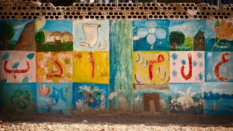 The image shows a wall decorated with colorful murals or paintings. The artwork includes various designs such as flowers, shapes, and possibly symbols or letters that are arranged in a grid pattern. The colors are vibrant, featuring shades of blue, green, yellow, and pink, giving the wall a lively and artistic feel. The surrounding area appears dusty or sandy, suggesting an outdoor environment. Overall, the scene conveys a sense of creativity and community.