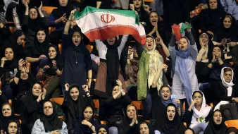 L'image montre un groupe de femmes dans une arène, probablement lors d'un événement sportif. Elles portent des vêtements traditionnels et modernes, et certaines d'entre elles agitent un drapeau iranien, exprimant leur soutien et leur enthousiasme. Les visages sont joyeux et animés, et l'atmosphère semble festive. Ce moment illustre la participation et la passion des femmes pour le sport en Iran.