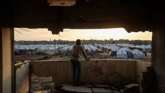 Un jeune observe un camp de tentes depuis une fenêtre endommagée au crépuscule.